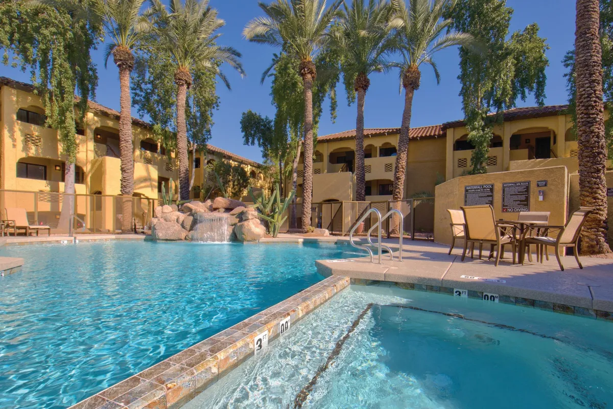 A sunlit resort pool area featuring a large swimming pool surrounded by palm trees, a connected jacuzzi hot tub, a cascading waterfall feature, and multiple lounge chairs set up for relaxation. The scene is lively yet serene, inviting guests to unwind or socialize by the water.