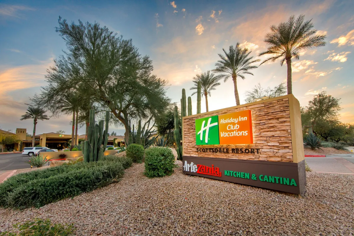 The main resort entrance, marked by a bold Holiday Inn Club Vacations sign, surrounded by lush landscaping, cacti, and tall palm trees. The welcoming setting hints at sunny weather and a vacation-ready environment, ideal for arrivals and outdoor exploration.