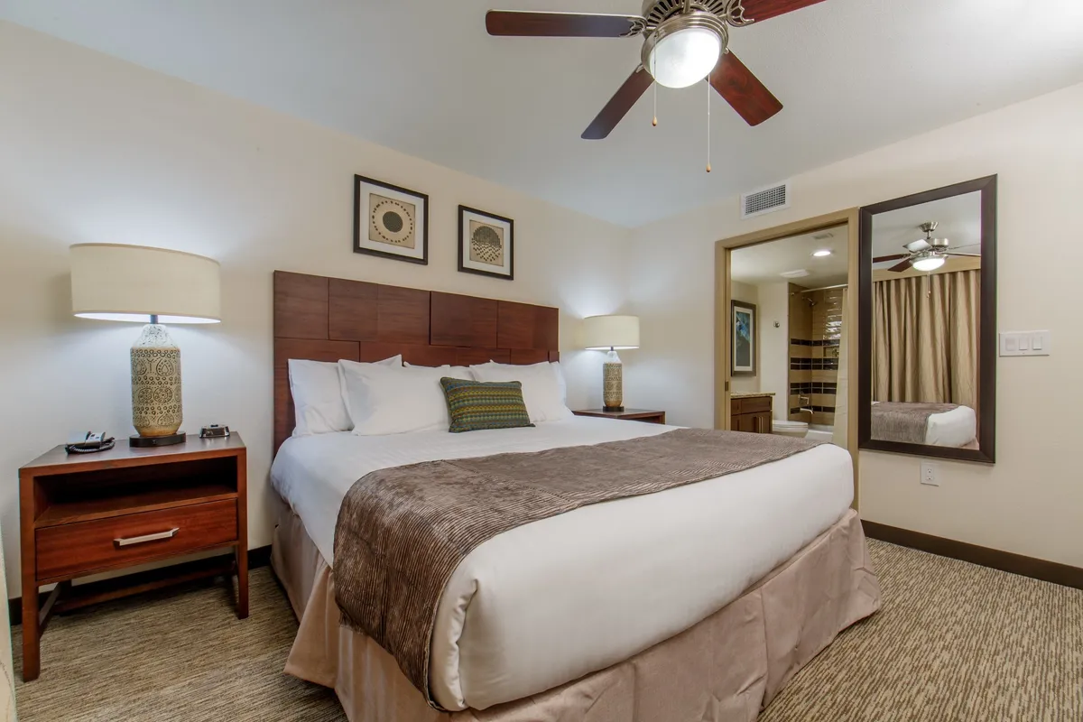 A spacious bedroom featuring a king-sized bed with plush linens, a nightstand with a warm lamp, decorative wall art that adds a personal touch, and a full-length mirror for convenience. The room is bathed in soft, natural light, creating an inviting and restful ambiance.