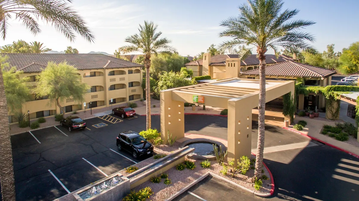 An inviting exterior view of the villa's entrance, featuring a canopy, a tranquil water feature, lush palm trees, and a convenient parking area. The setting feels peaceful and welcoming, ideal for guests arriving for a relaxing stay.