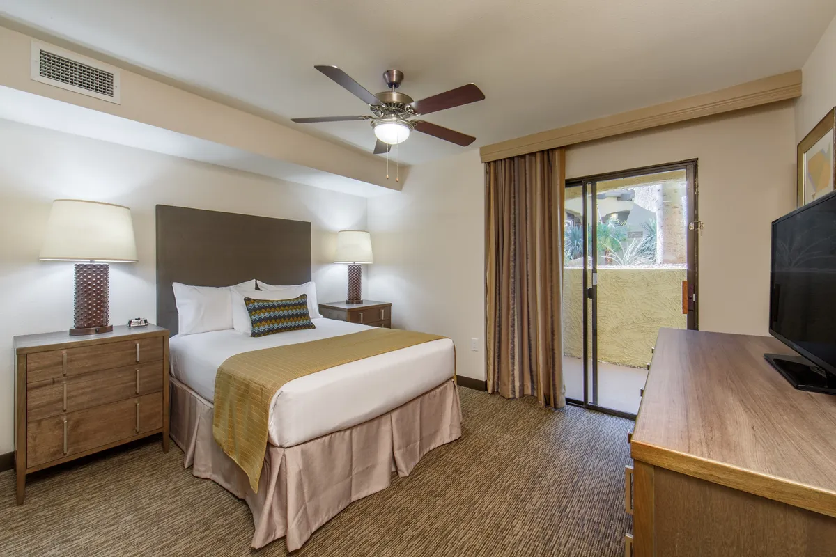 This image captures a cozy, well-appointed bedroom featuring a plush bed centered between nightstands, a TV on the wall, and a balcony door that allows natural light to pour in. The open door hints at a private outdoor space beyond, offering a seamless connection between the villa’s interior and the lively world outside.
