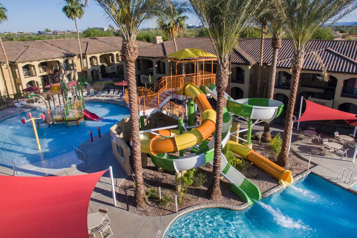 A vibrant outdoor swimming pool area featuring twisting water slides, tall palm trees, and ample shaded seating. The pool is lively, with spaces for families to gather, splash, and relax. The water slides are a focal point, inviting kids and adults alike for thrilling fun.